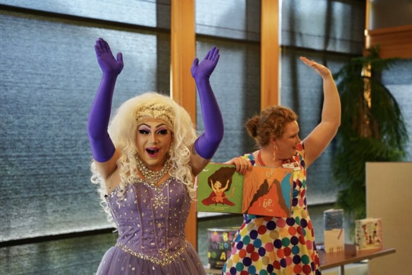 A woman in a platinum blond wig and purple princess dress raises her arms in the air with above-the-elbow purple gloves on